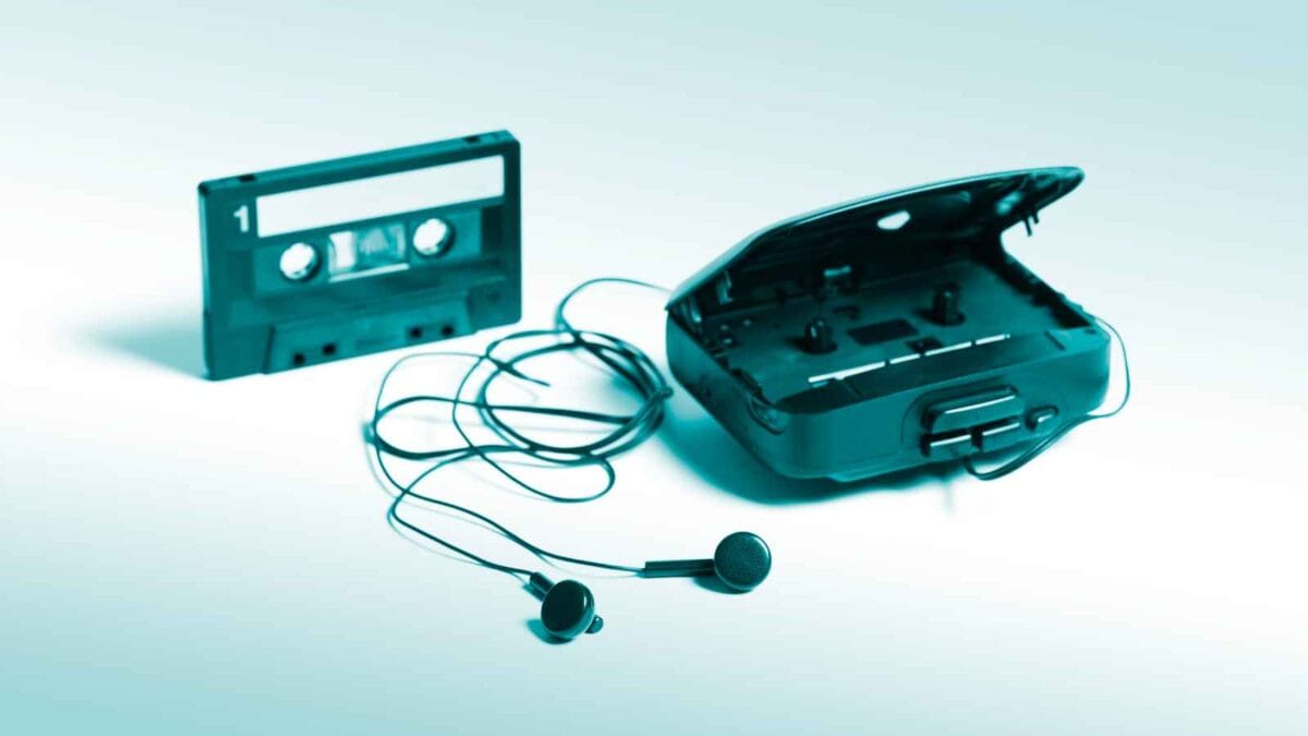 An old cassette tape beside a portable cassette player with its lid open and black earphones on a light blue background, representing things-that-were-once-great-in-our-society-but-are