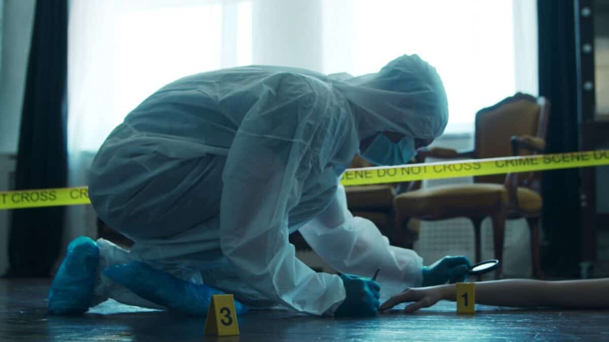 A forensic investigator in a protective suit examines a clue next to a body at an indoor crime scene with 'CRIME SCENE DO NOT CROSS' tape in the foreground.