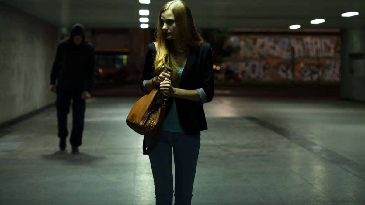 A cautious woman walking in an underpass with a hooded figure in the background, in a dimly lit urban environment