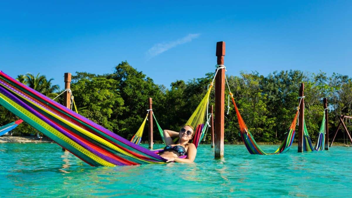 A person relaxes in a colorful hammock suspended over crystal-clear turquoise water, with lush greenery in the background under a blue sky.