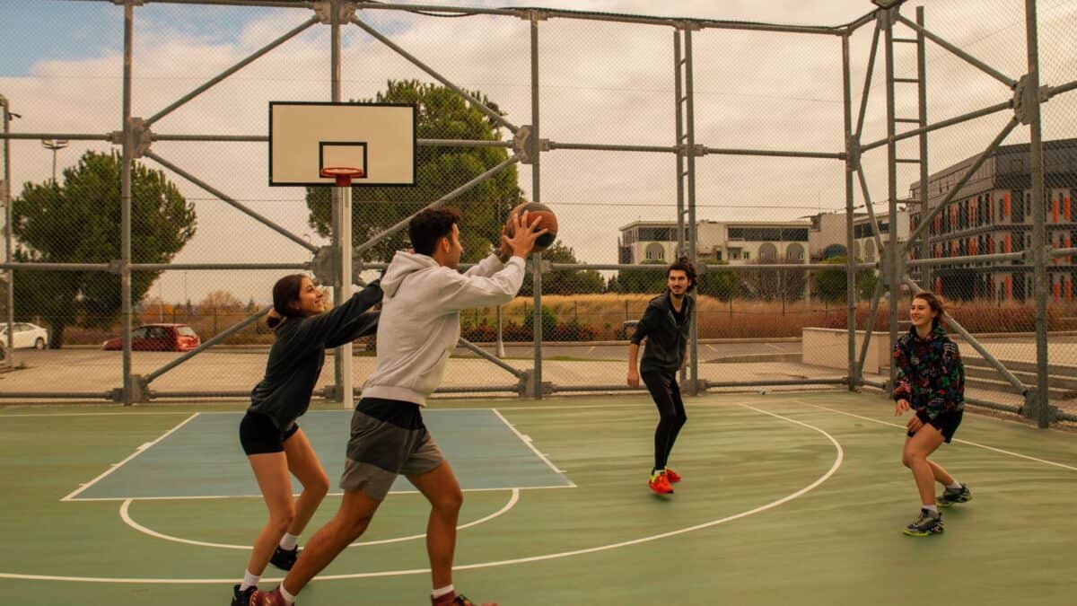 Four young adults playing basketball on an outdoor court, with one person leaping to shoot the ball as others prepare to defend or catch.