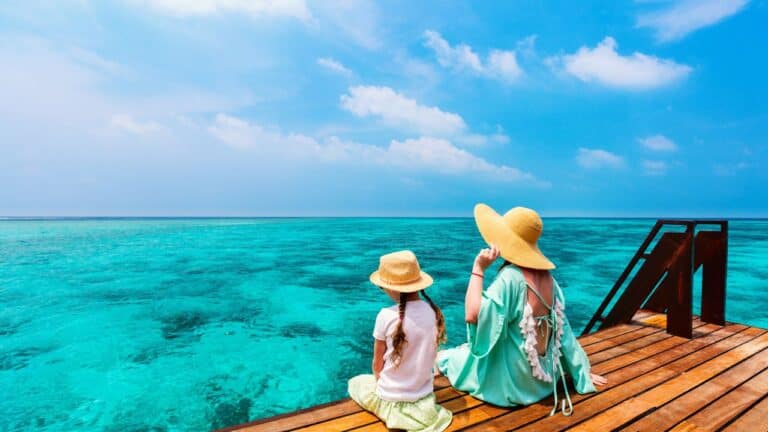 Two individuals, an adult and a child, both wearing wide-brimmed hats, sitting on a wooden dock looking out at the vibrant blue ocean.