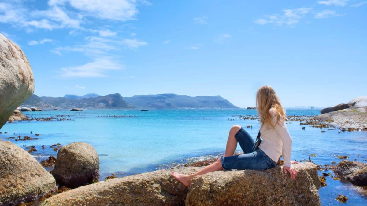Blonde woman sitting on a boulder by the sea, with a panoramic view of a coastal landscape and mountains in the distance."