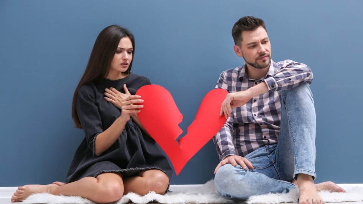 A man and a woman sit on a white rug against a blue wall, looking sad and holding two halves of a broken red heart. the atmosphere is somber, reflecting emotional distress.