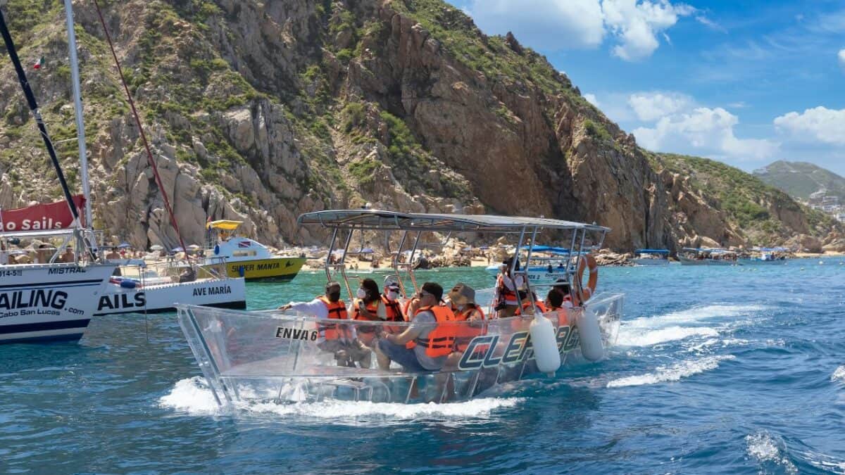 Tourists in orange life vests enjoy a boat tour near rocky cliffs on a sunny day in Cabo San Lucas, with other boats in the background.