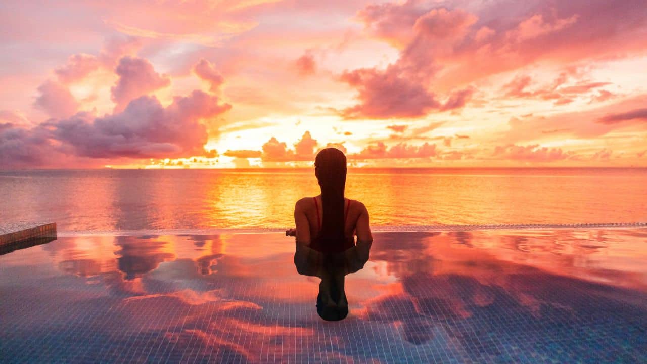 A person meditating in an infinity pool against a vibrant sunset with orange and pink clouds reflected in the water.