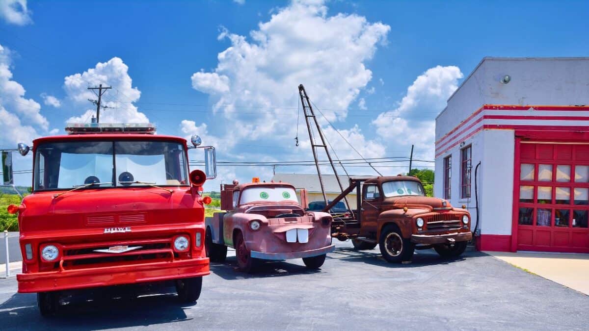 Three vintage trucks with anthropomorphic features displayed outside a classic service station under a blue sky with fluffy clouds.