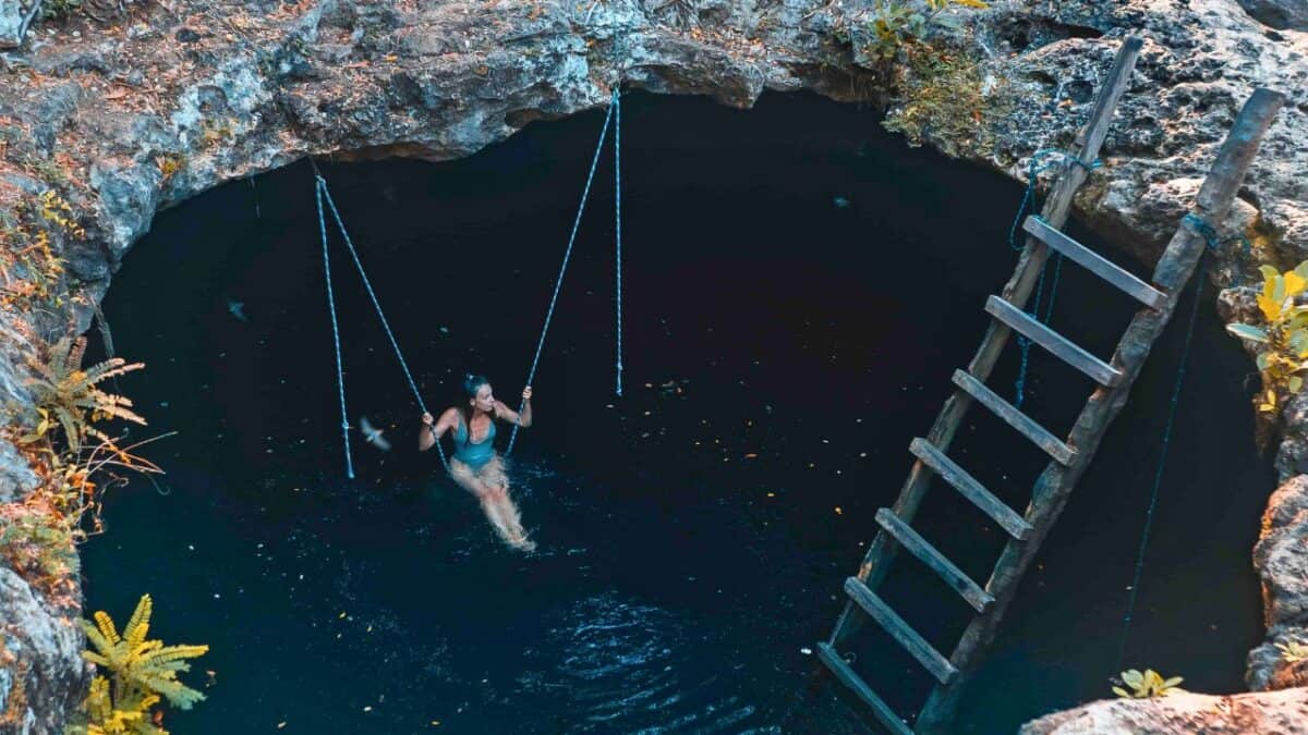 A person swinging on a rope above a clear natural water hole surrounded by rocky terrain and lush vegetation.