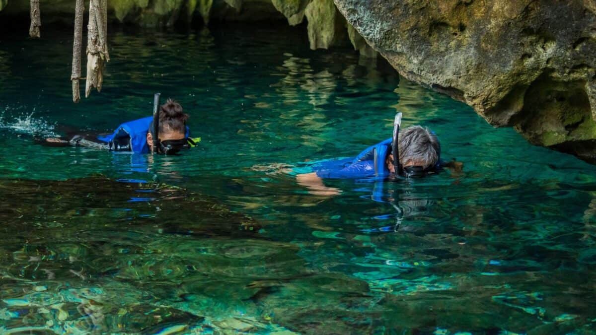Two people snorkeling in clear, turquoise water near the rocky edges of a cenote.