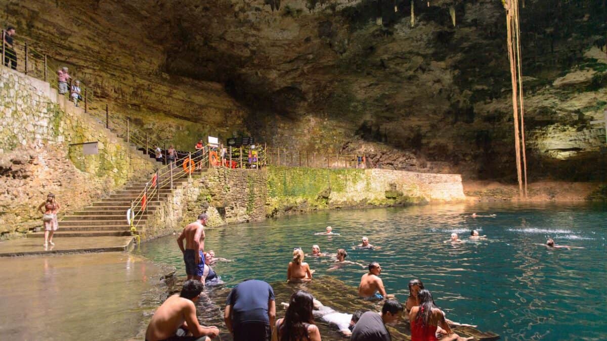 Tourists swimming and relaxing in the clear waters of the Hubiku Cenote, an underground natural pool with a dramatic rock ceiling and hanging roots.