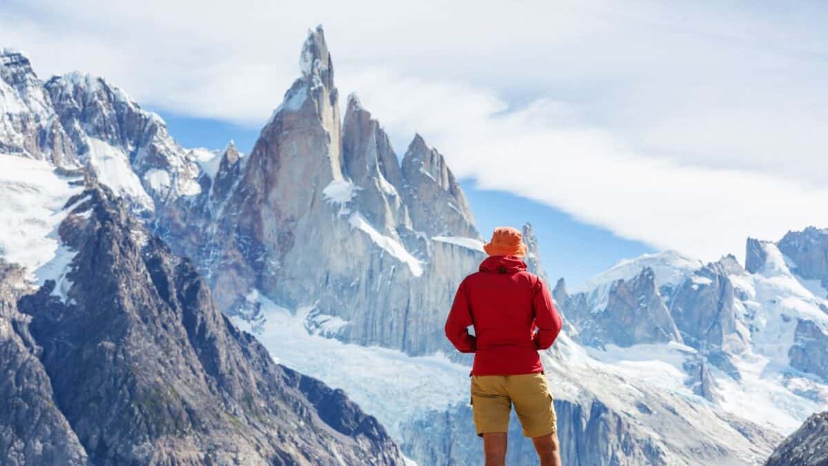 A person in a red jacket and orange hat standing and looking at the jagged peaks of Cerro Torre with glaciers visible in the background.