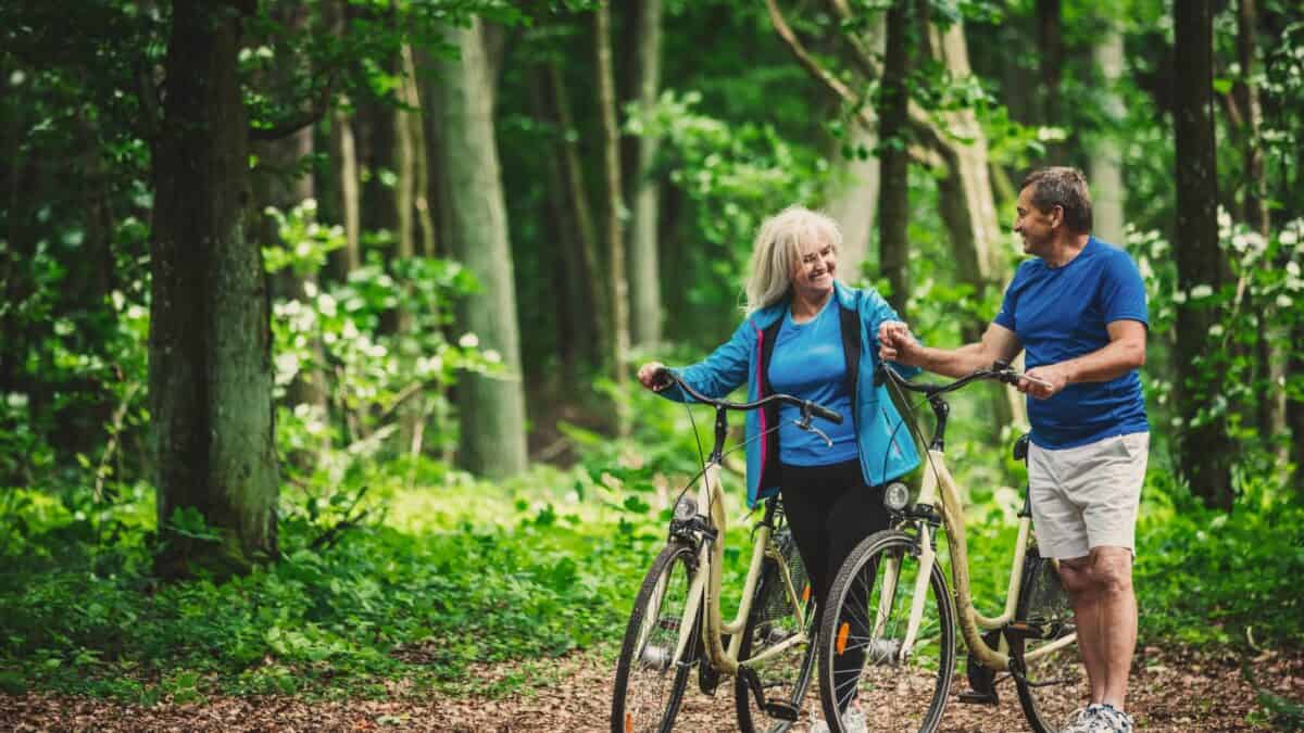 A senior couple with bicycles in a lush green forest, smiling and chatting while standing on a dirt path. the woman is blonde, and they are both wearing casual blue outfits.
