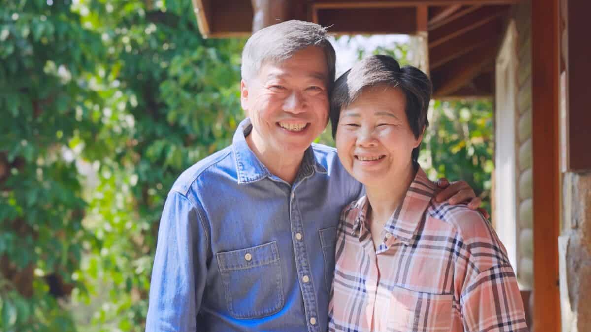 An older asian couple smiling warmly, standing together under a sunlit porch, surrounded by greenery. the man wears a denim jacket and the woman a plaid shirt.