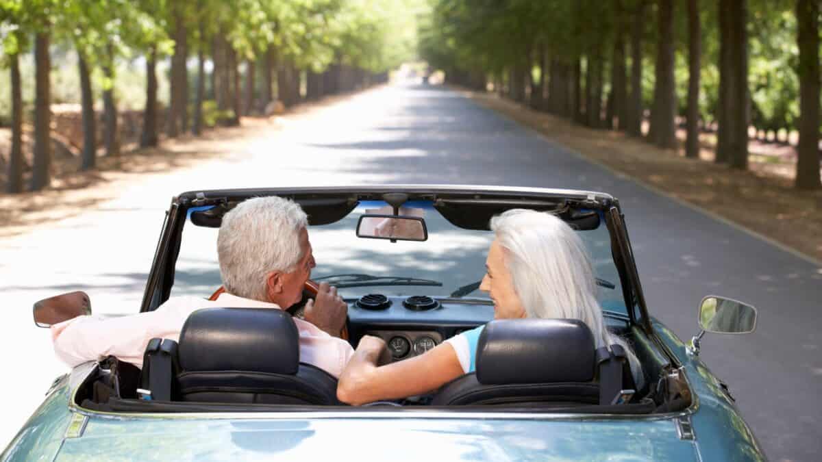 An elderly couple enjoys a summer drive in a convertible car along a tree-lined road, viewed from behind.