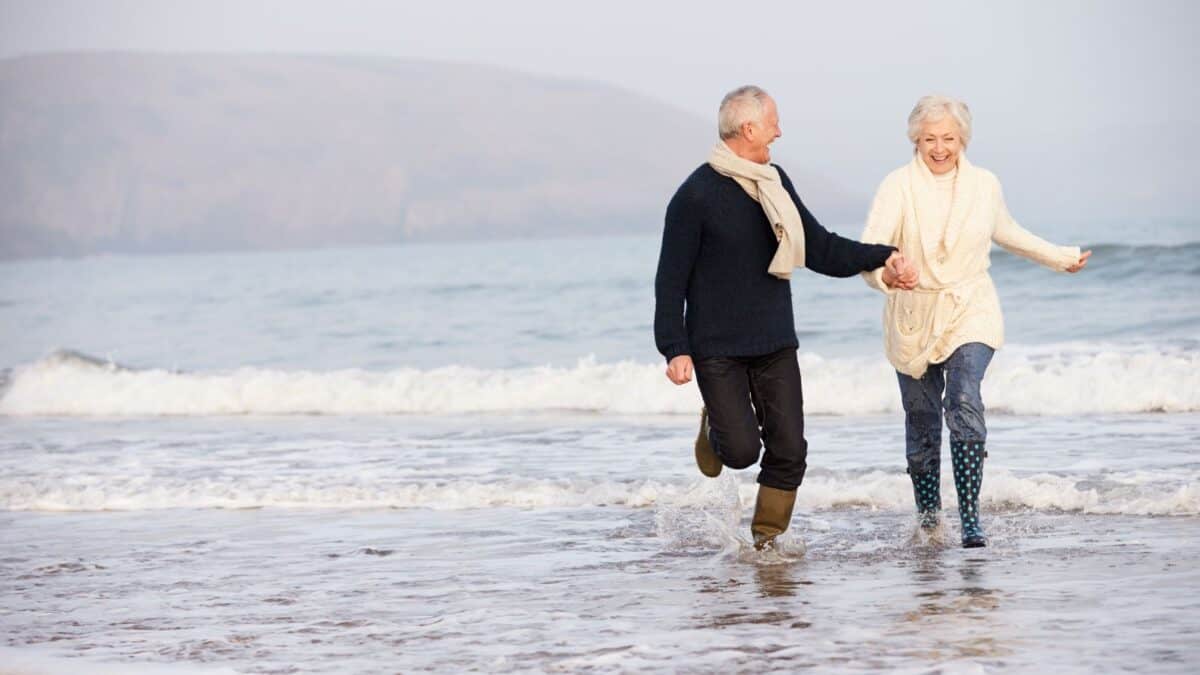 An elderly couple happily holding hands and running along a beach, with gentle waves at their feet and a soft, misty hill in the background.