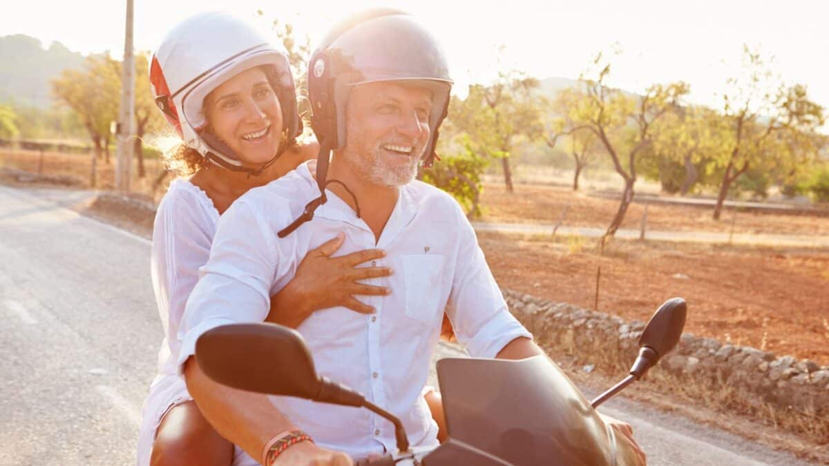 A joyful older man and a young woman wearing helmets riding a scooter on a sunny rural road, with trees in the background. the woman is embracing the man from behind with a smile.