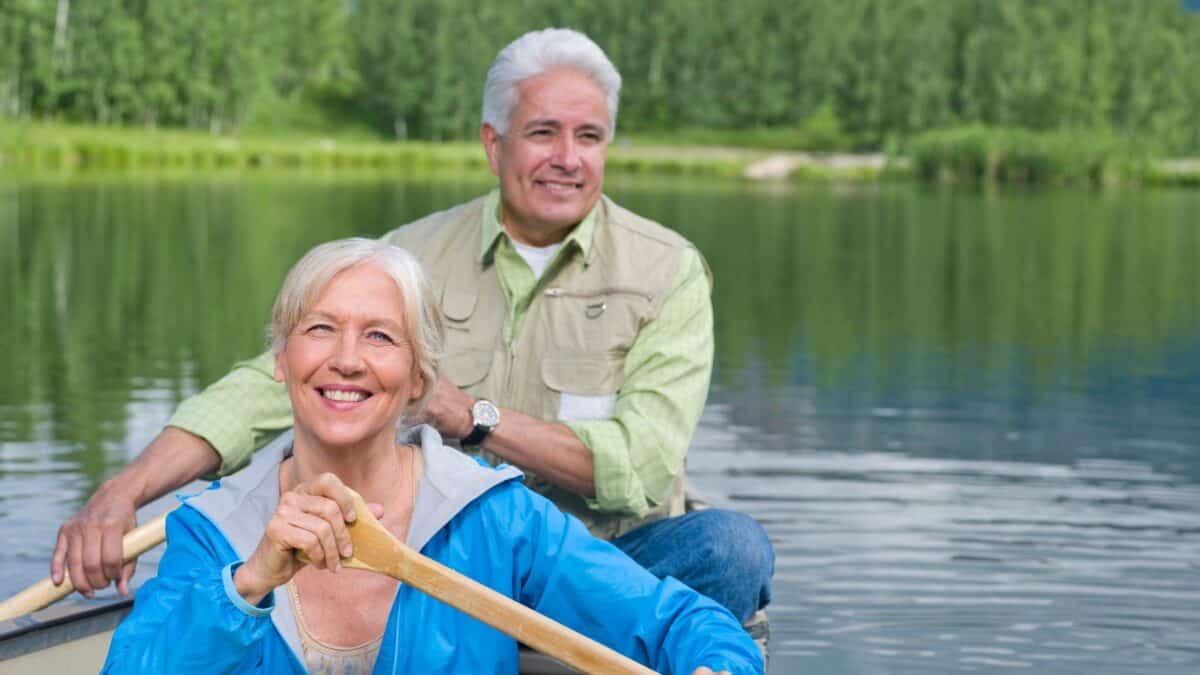 An elderly couple in a canoe on a tranquil lake, with the woman in front smiling at the camera and the man seated behind her, both enjoying the day.