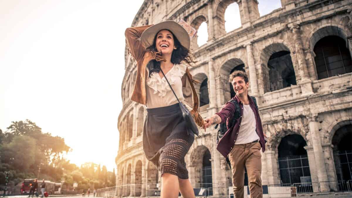 A joyful couple holding hands and running in front of the colosseum in rome, with the woman leading and looking back at her partner, both smiling brightly.