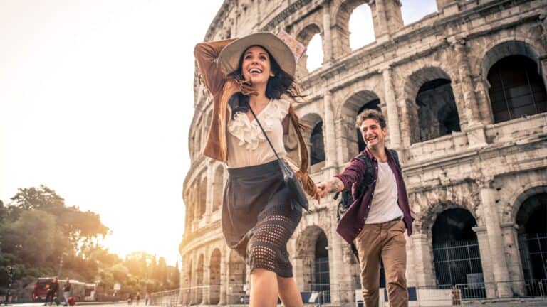 A joyful couple holding hands and running in front of the colosseum in rome, with the woman leading and looking back at her partner, both smiling brightly.