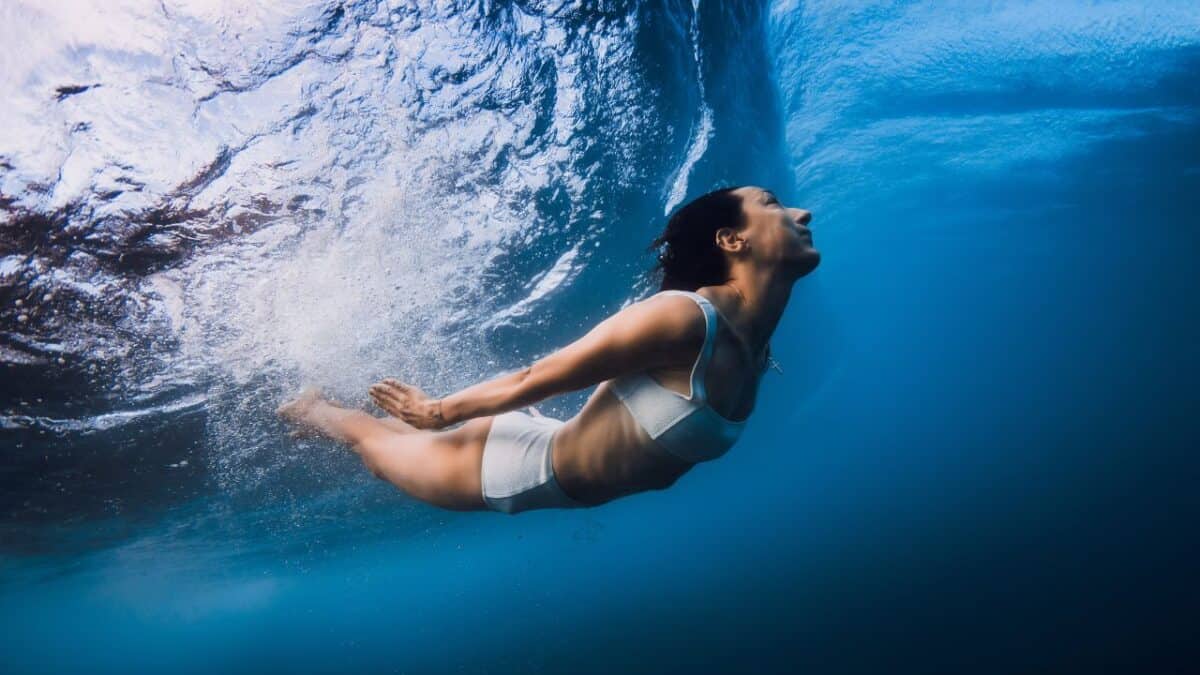 A woman in swimwear gracefully free diving underwater, with light refracting on the surface above her.