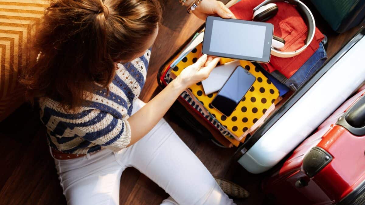 A woman packing a suitcase with clothes and gadgets, holding a smartphone with a blank screen, viewed from above.