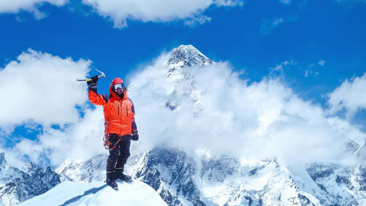 A mountaineer in an orange jacket standing victoriously on a snowy peak with an ice axe raised in hand, with the cloud-shrouded summit of Mount Everest in the background.