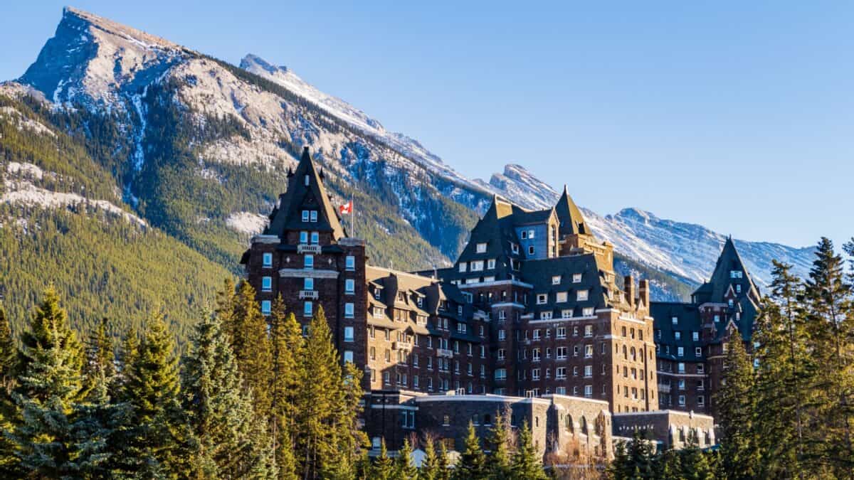 A historic castle-style hotel with multiple steep roofs and towers, nestled at the base of a large, snow-capped mountain range under a clear blue sky.