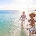 Two joyful children, one with curly hair, playfully running towards the camera on a sunlit beach with clear blue water and a distant pier.