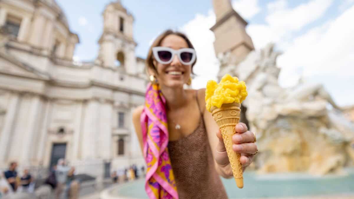 20 Phrases You Should Learn Before You Visit Italy 6 A joyful woman in sunglasses and a pink scarf holds out a gelato cone with a blurry historic fountain and buildings in the background.