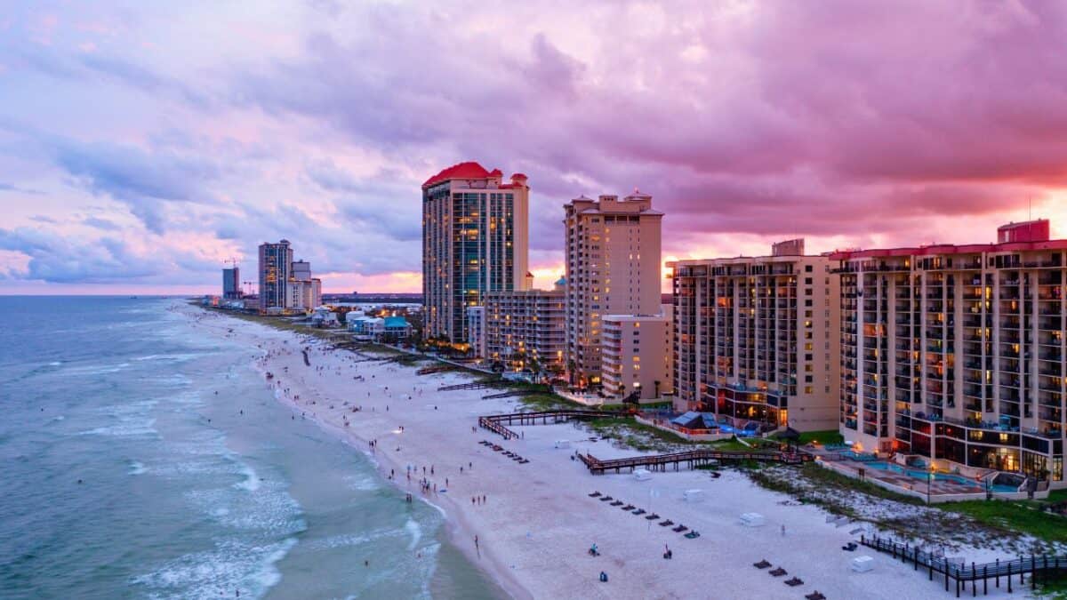 Aerial view of a beach at sunset with high-rise buildings along the shore, vibrant pink and orange sky, and people on the sandy beach.