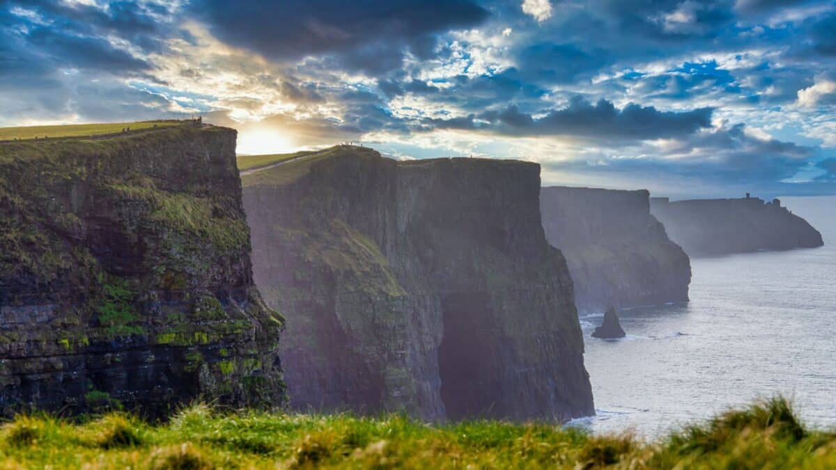 Sunset over the cliffs of moher in ireland, with rays of sunlight breaking through the clouds above the rugged green cliffs that descend sharply into the atlantic ocean.