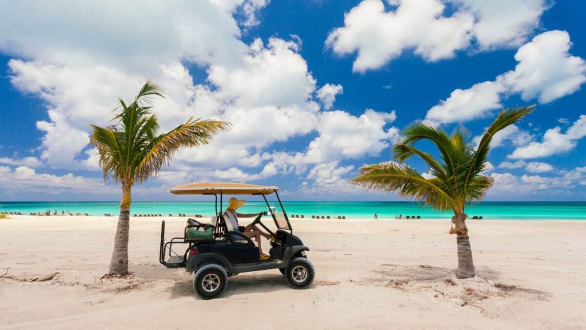 A person in a wide-brimmed hat drives a golf cart on a serene beach flanked by palm trees, with the turquoise ocean and a clear blue sky in the background.