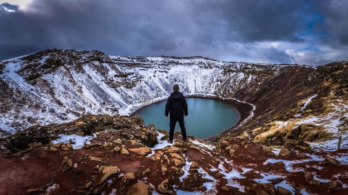 A person stands at the edge of Kerið volcanic crater, looking at the snow-dusted slopes and the vivid blue water at the bottom.