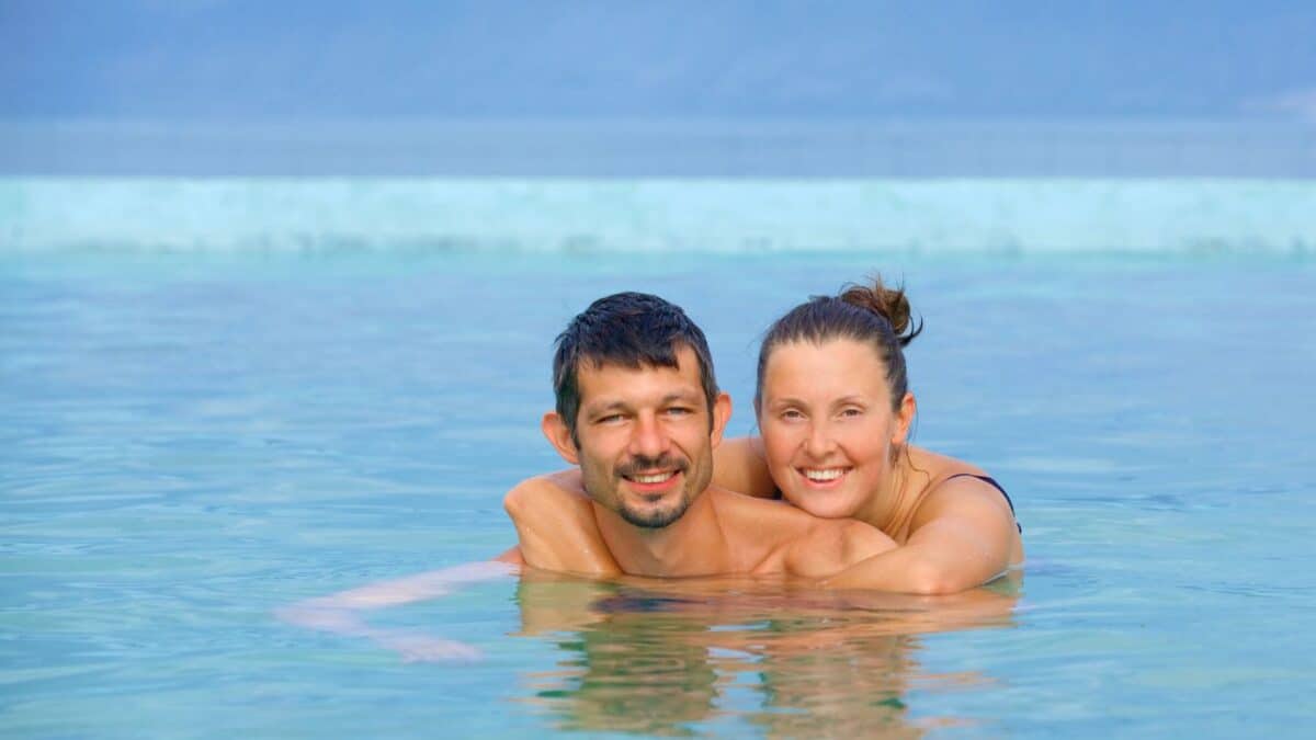 A couple smiling while embracing in the blue waters of a geothermal spa with mountains in the background.