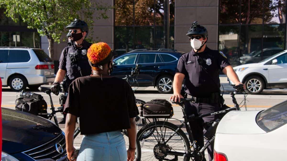 A civilian with colorful hair facing two bike patrol officers in a city parking scene with cars and trees in the background.