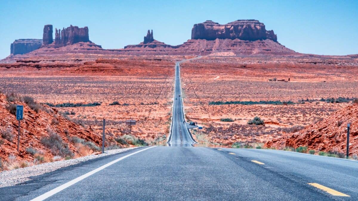 A straight highway extends into the distance toward large red rock formations under a clear blue sky in a desert landscape.