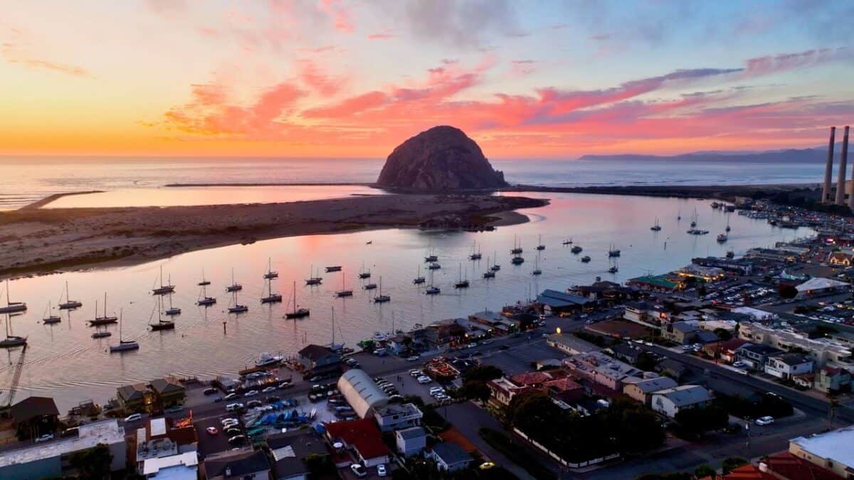 Aerial view of a harbor at dusk with moored boats, a distinctive rocky hill in the background, and a small town with glowing lights under a sky with hues of pink and orange.