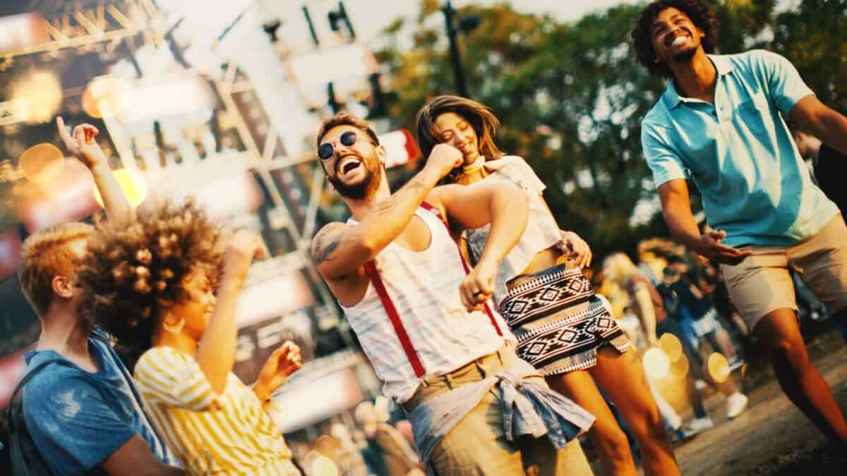 A group of young adults joyfully dancing at an outdoor festival, with warm sunlight filtering through trees in the background.