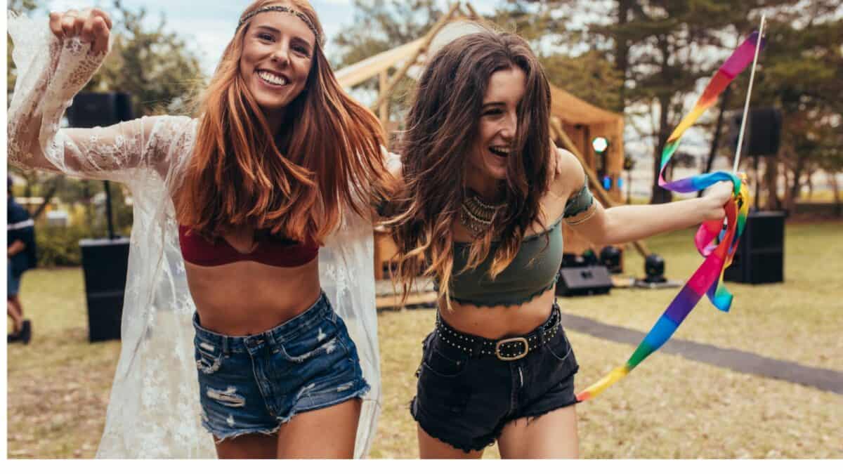 Two joyful young women dancing and waving ribbons at an outdoor festival, surrounded by trees and a tent in the background. they are smiling and enjoying the moment.