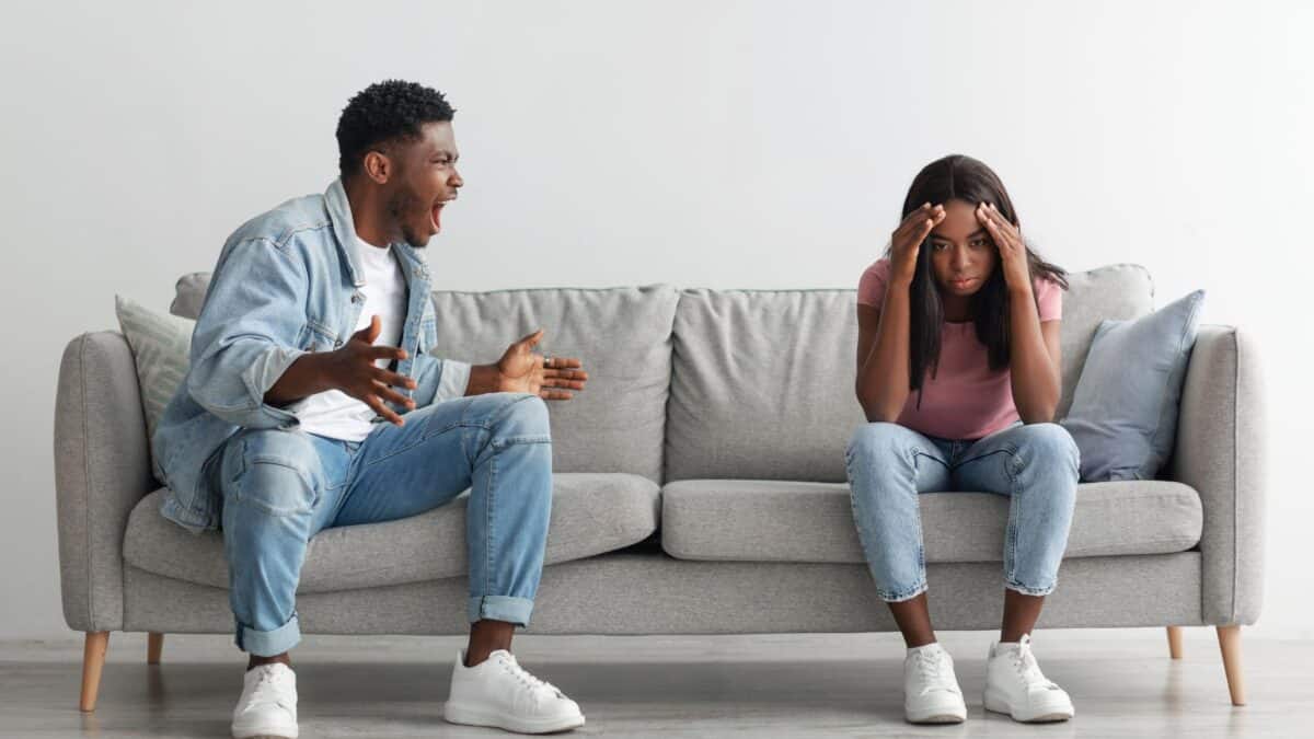 A man and a woman sitting on a gray sofa in a bright room, having a conversation where the man is gesturing animatedly while the woman appears upset, with her head in her hands.