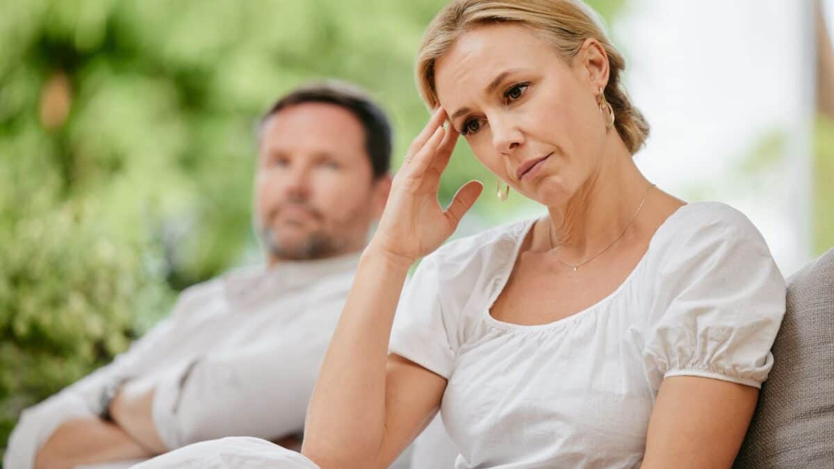 A woman looks distressed, touching her forehead, sitting next to a man who appears contemplative. both are seated on a couch in a garden setting, indicating some tension or disagreement.