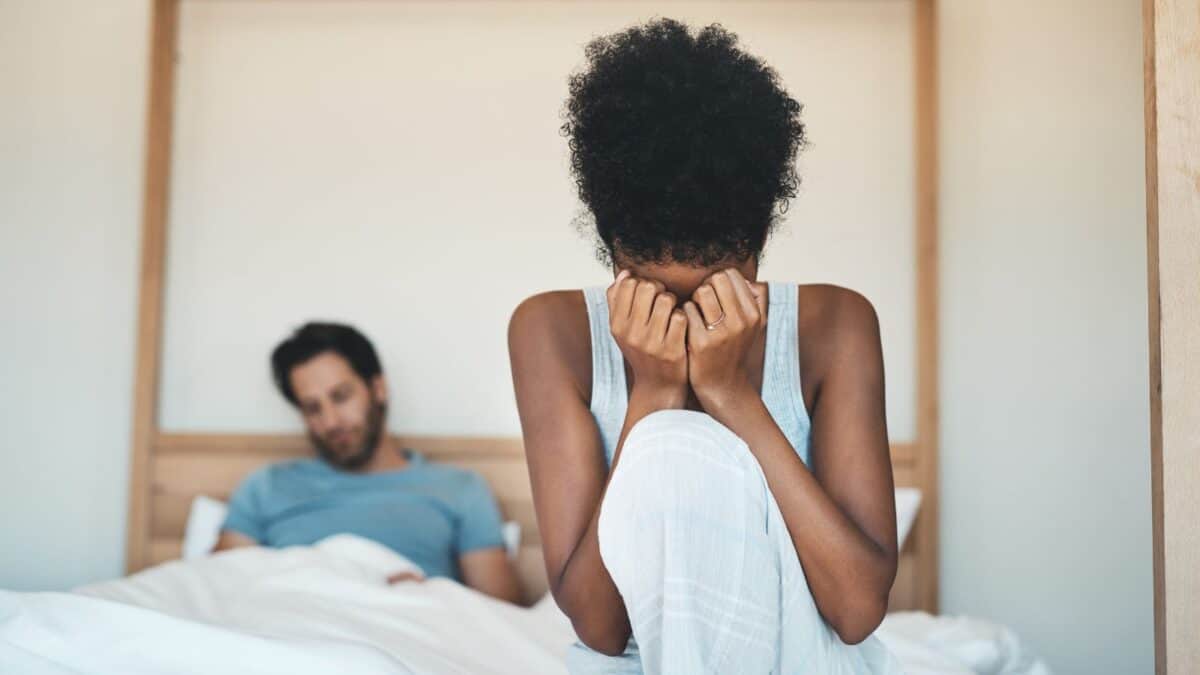 Inside the Narcissist's Playbook: 20 Tactics to Control and Confuse 15 A distressed woman sitting on the edge of a bed, covering her face with her hands, while a man sits in the background, watching her with a concerned expression.