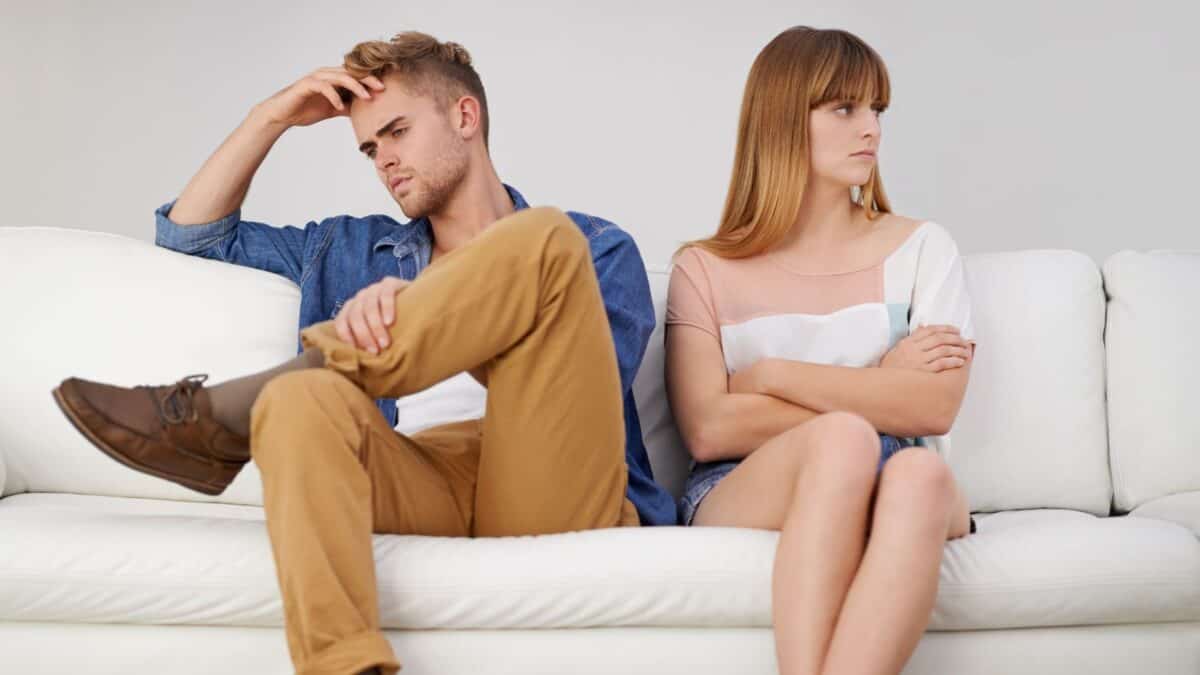A young couple sitting on a white couch appears upset with each other, avoiding eye contact, with arms crossed and a troubled expression on their faces.