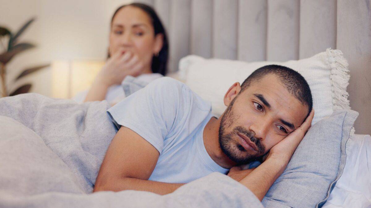 Inside the Narcissist's Playbook: 20 Tactics to Control and Confuse 7 A man with a beard appears upset, lying on a bed with his head on his hand, while a woman in the background looks thoughtful, both appearing emotionally distant.