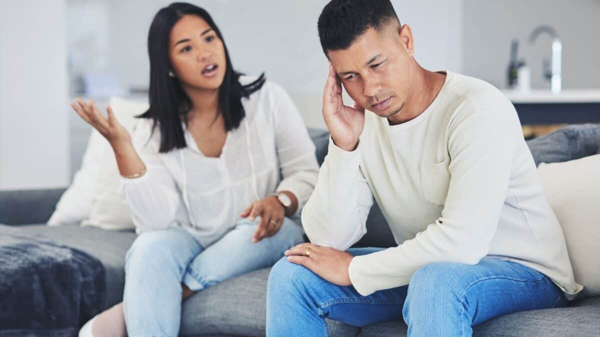Inside the Narcissist's Playbook: 20 Tactics to Control and Confuse 8 A man, looking frustrated, sits with his head in his hand on a sofa, while a woman gestures animatedly, appearing to argue with him, in a modern living room.