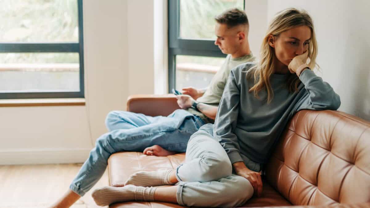 Inside the Narcissist's Playbook: 20 Tactics to Control and Confuse 6 A young couple sitting on a couch in tension, not facing each other; the woman appears upset, leaning her head on her hand, while the man looks away with a phone in hand.