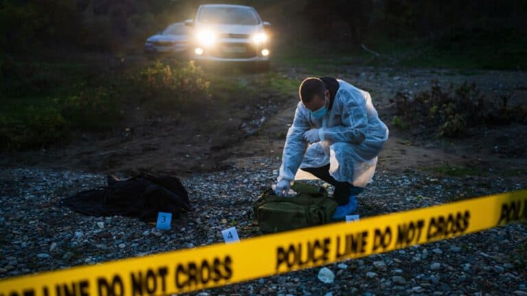 Forensic expert in protective clothing examining evidence at an outdoor crime scene with police tape in the foreground and a patrol car in the background at dusk.