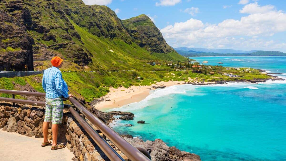 15 Best Cities In The United States For Thanksgiving Celebrations 12 A person in a bright orange hat and casual attire overlooks a stunning beach with turquoise waters and lush green mountains in the background.