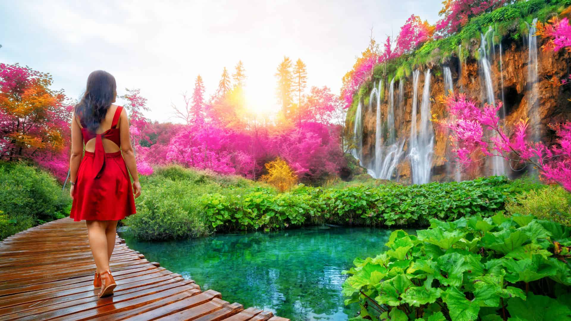 A woman in a red dress walking on a wooden boardwalk towards a vibrant, multicolored waterfall surrounded by lush foliage.