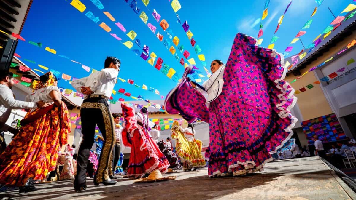 Dancers in traditional Mexican attire perform a folk dance under colorful papel picado streamers, with bright blue sky above.